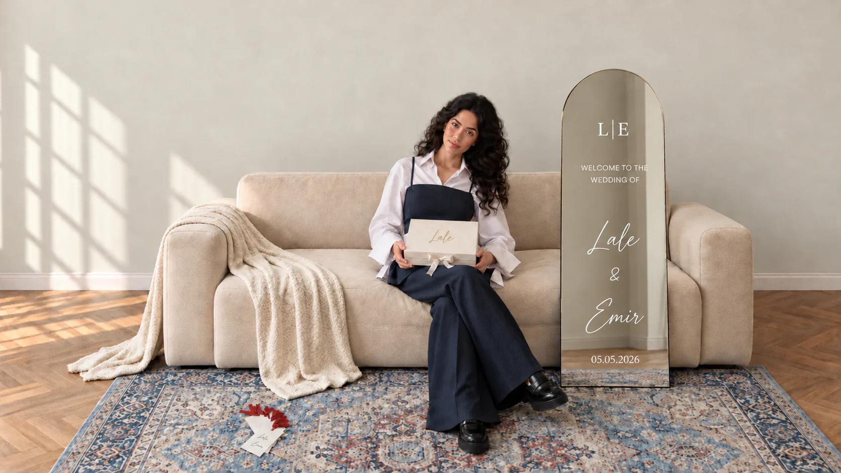 Frau auf einem beigen Sofa mit einer personalisierten DUVAM-Geschenkbox mit dem Namen Lale im Schoß; daneben ein bogenförmiger Standspiegel mit weißer Klebeschrift „Welcome to the wedding of Lale & Emir, 05.05.2026“