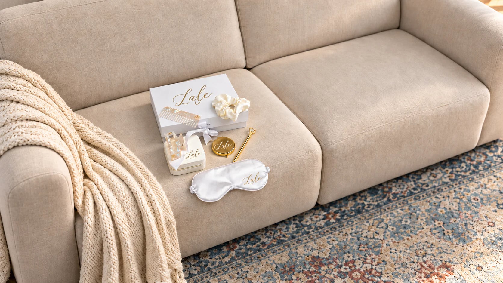 Personalisiertes Trauzeugin-Set auf einem beigen Sofa: weiße Box, Schmuckdose, Haarkamm, Scrunchie, goldener Taschenspiegel, Stift und Schlafmaske aus Satin, jedes Stück mit dem Namen Lale in Gold