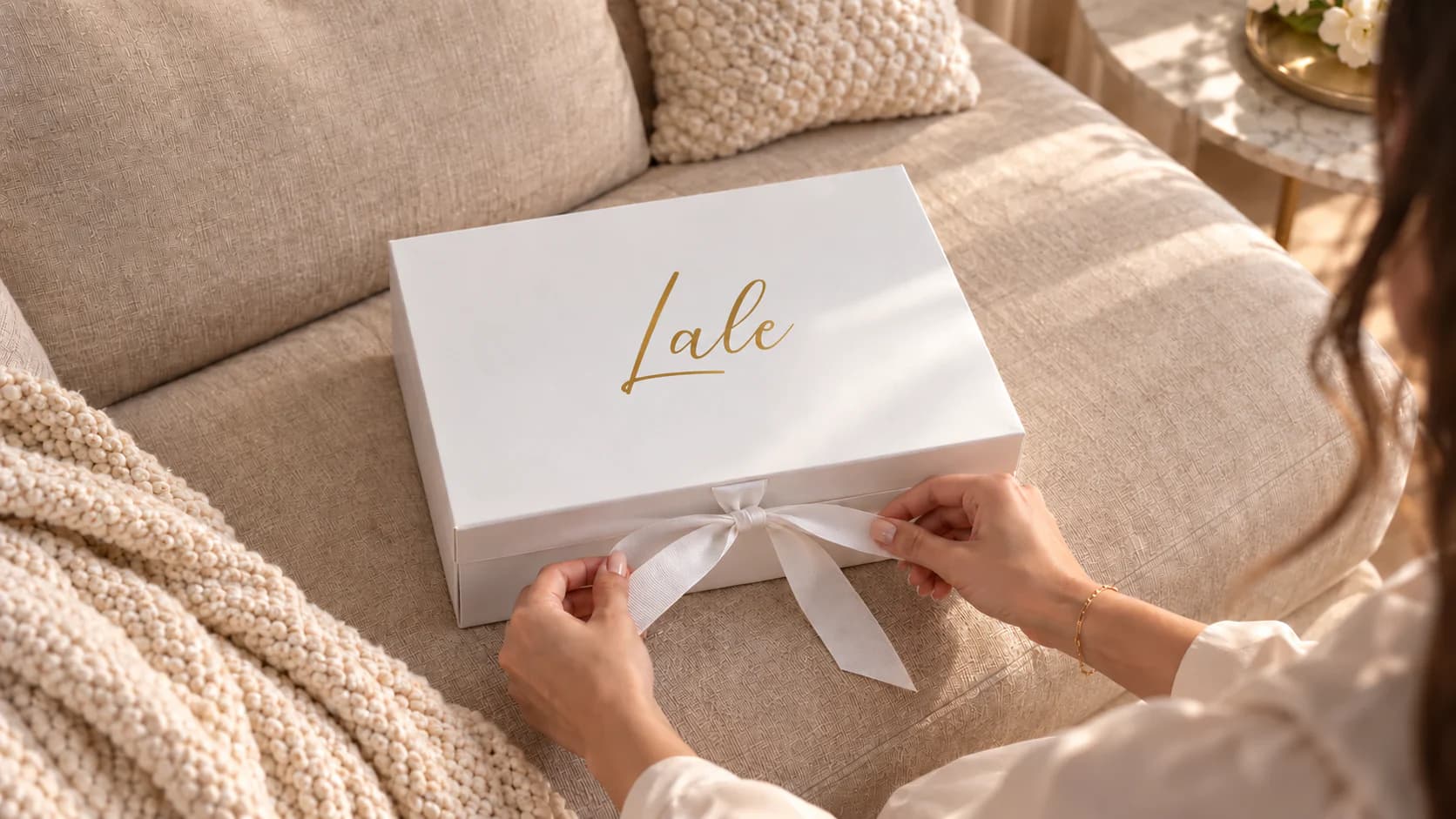 A white bridesmaid gift box with a gold name on a beige sofa; hands tie its white ribbon.
